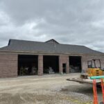 Installed shingle roof above three open garage bays in a brick addition, with construction equipment in front.
