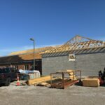 Exposed wood roof trusses above a concrete-block addition beside an existing brick building with partially exposed roof decking.