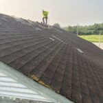 Worker removing existing shingles from a sloped roof above a weathered green-trimmed gable.