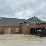 Intersecting shingle roof sections above brick walls, with boarded openings and construction dumpsters below.